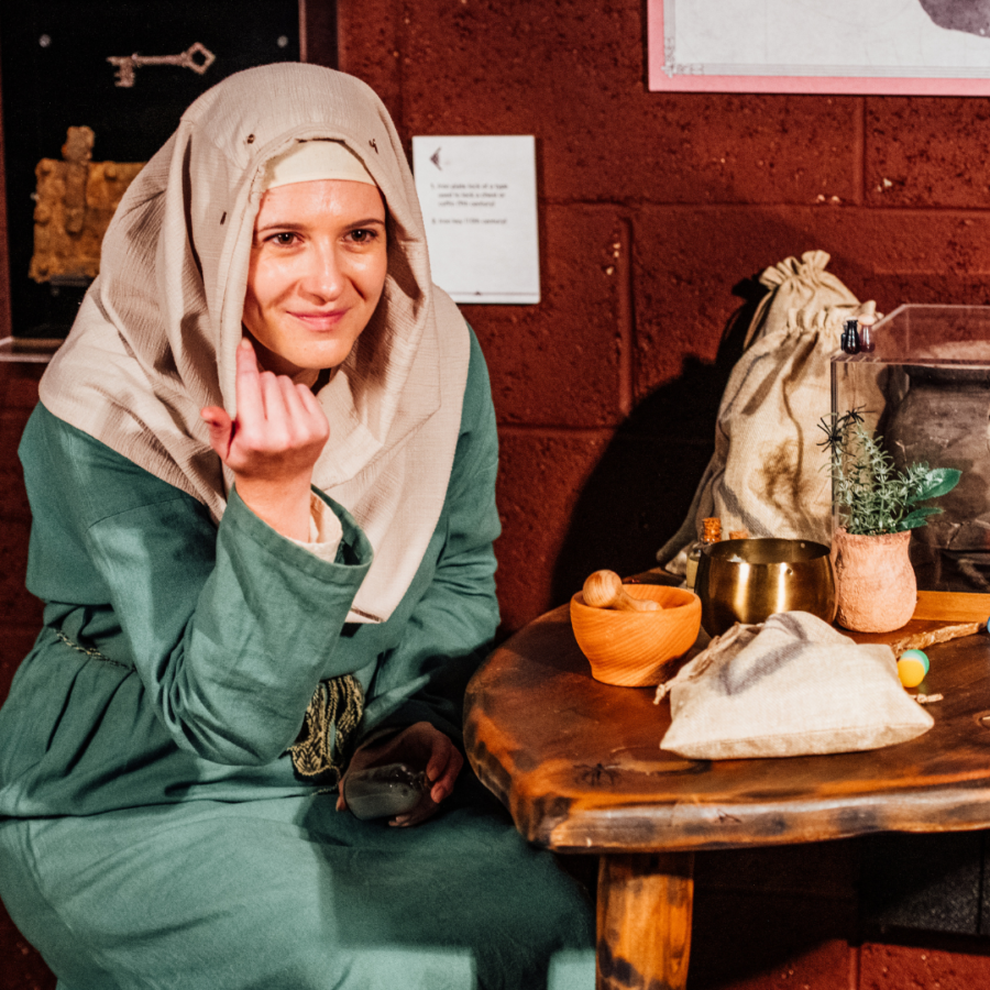 A woman in Anglo Saxon attire, next to a wooden table entices you to join her