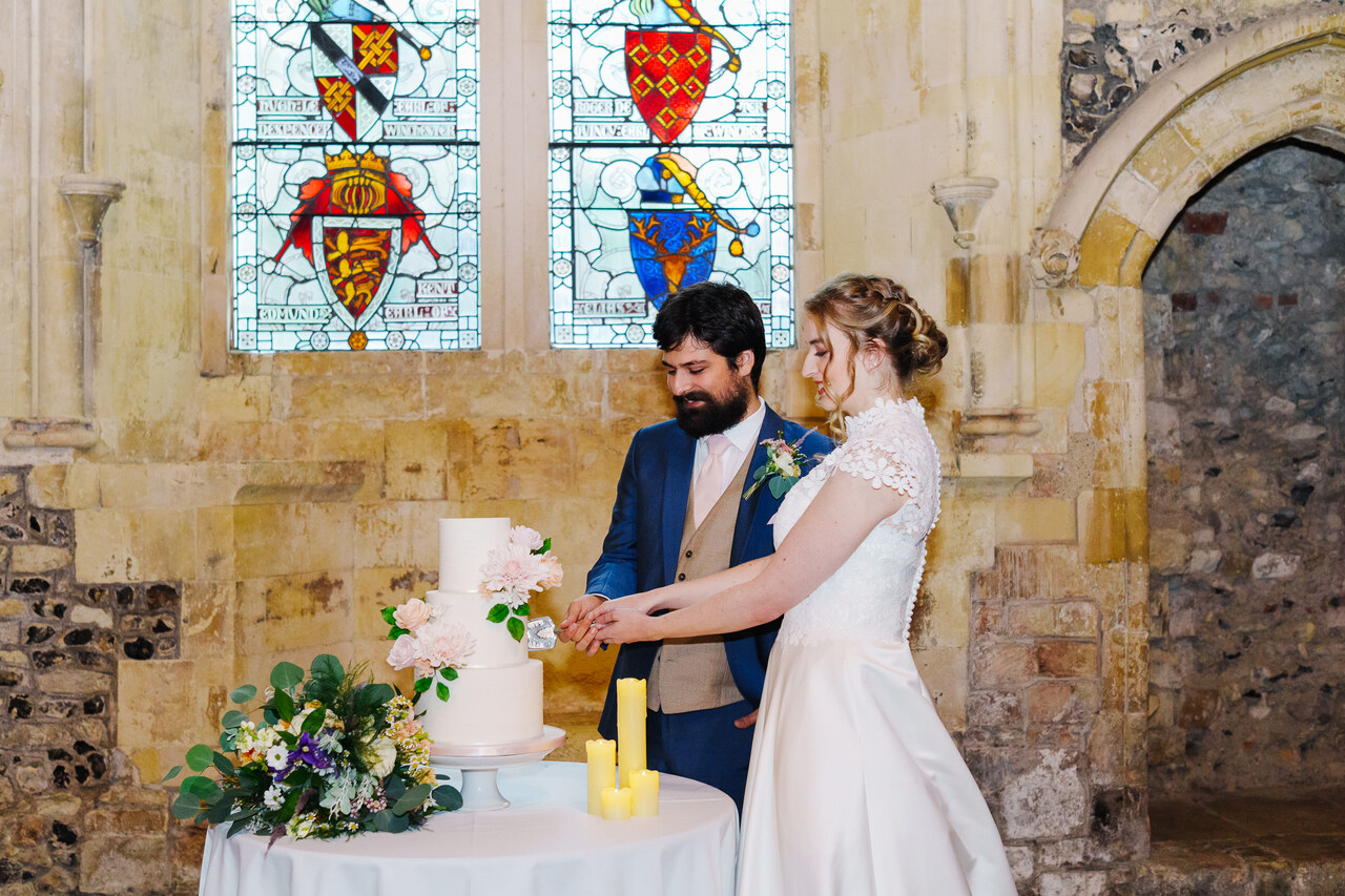 Two people in wedding attire cut a cake Infront of an old wall