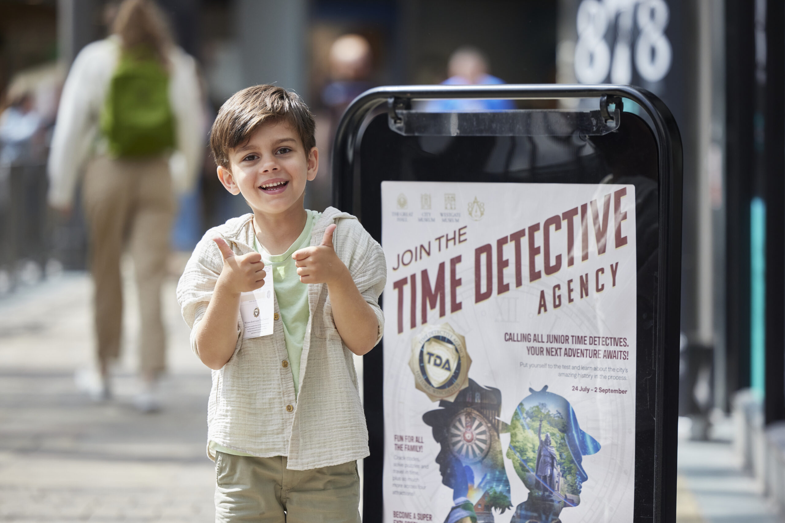 A boy with his thumbs up in front of a Time Detective Agency sign.